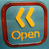 Sobre superficie metálica, botón con bordes redondeados y en su interior una señal en la que se puede leer la palabra “open” y dos símbolos indicativos hacia la izquierda.