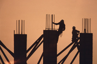 Fotografía a contraluz en la que se ven las siluetas de dos operarios trabajando en la construcción de vigas.