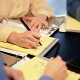 Sobre una mesa de oficina, se pueden ver las manos de dos técnicos realizando anotaciones en sus blocs de hojas amarillas.
