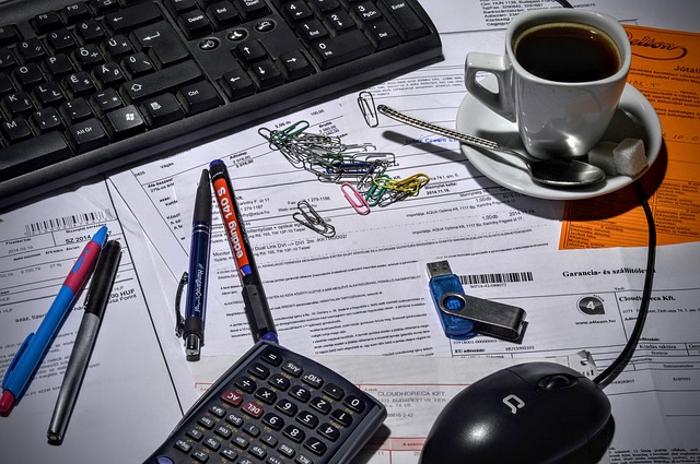 Foto de una mesa llena de documentos dispersos, sobre los que se ve un teclado de ordenador, unos clips, unos bolígrafos y una taza de café.