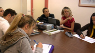  Imagen de cinco personas sentadas en una mesa, donde hay un proyector funcionando y una mujer explicando algo, mirando hacia una pantalla.