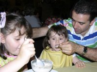Imagen que muestra a un hombre y a sus dos hijas. Una de las hijas está comiendo por sí sola, pero la otra es demasiado pequeña. La imagen muestra al padre dándole de comer a la niña. 