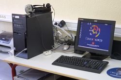 En una mesa aparece un ordenador, monitor, impresora y cámara web.