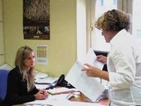 Foto de dos compañeras de trabajo en una oficina, una de ellas es Elena que está sentada hablando con Carmen que está de pie con papeles en la mano.
