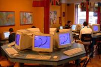 Sala de ordenadores donde se ve en un primer plano 3 ordenadores encendidos pero vacíos y al fondo gente trabajando con otros ordenadores.
