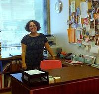 Una mujer sonriente, con gafas, está de pie junto a su mesa de trabajo