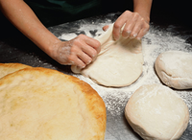 En una mesa de un obrador de pan, las manos del panadero amasan la harina para darle forma. Al lado, un pan ya cocido y dos hogazas aún por cocer.