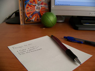 Papel y bolígrafos sobre una mesa de escritorio, al fondo la parte de abajo de un monitor una pequeña pelota de color verde.