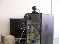 Muestra la cámara web situada sobre la caja del ordenador y conectada a un conector tipo USB en la trasera del ordenador. También se puede ver el resto de periféricos ya conectados.